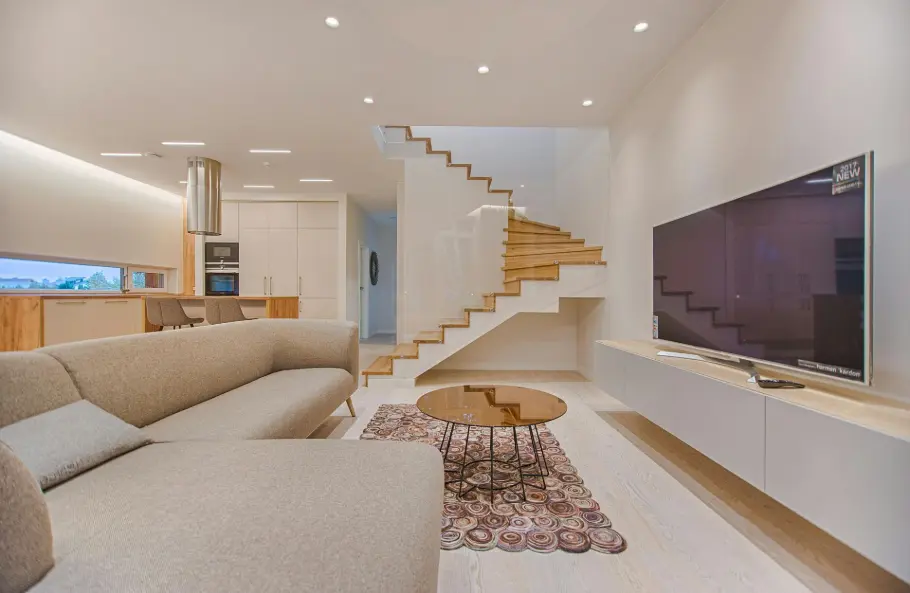 Un intérieur de maison moderne et minimaliste avec un canapé beige confortable, une cuisine ouverte, un escalier en bois et un téléviseur élégant. Représente le rôle des diffuseurs nébulisants dans l'amélioration de la qualité de l'air intérieur, de la relaxation et du bien-être général à domicile.