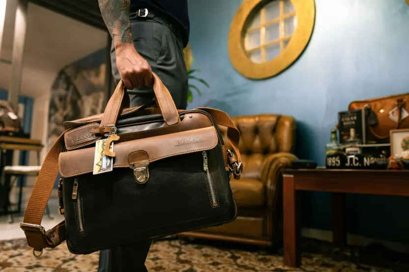 Un sac de bureau en cuir élégant avec des boucles en laiton posé sur un bureau dans un espace de travail professionnel, avec un fauteuil en cuir vintage et une décoration de bureau. Représente la productivité, la concentration et le bien-être au travail, en accord avec les bienfaits de la diffusion d'huiles essentielles au bureau.