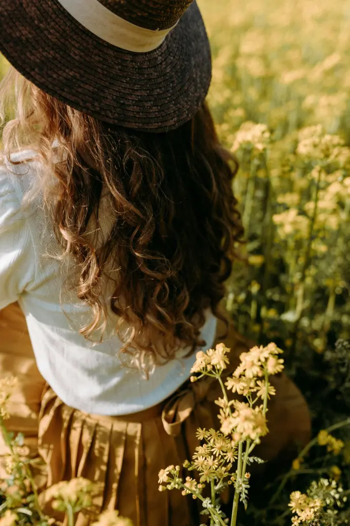 A woman with curly hair wearing a straw hat and a white blouse, standing in a sunlit field of yellow flowers. Represents nature, wellness, and the essence of botanical aromatherapy.